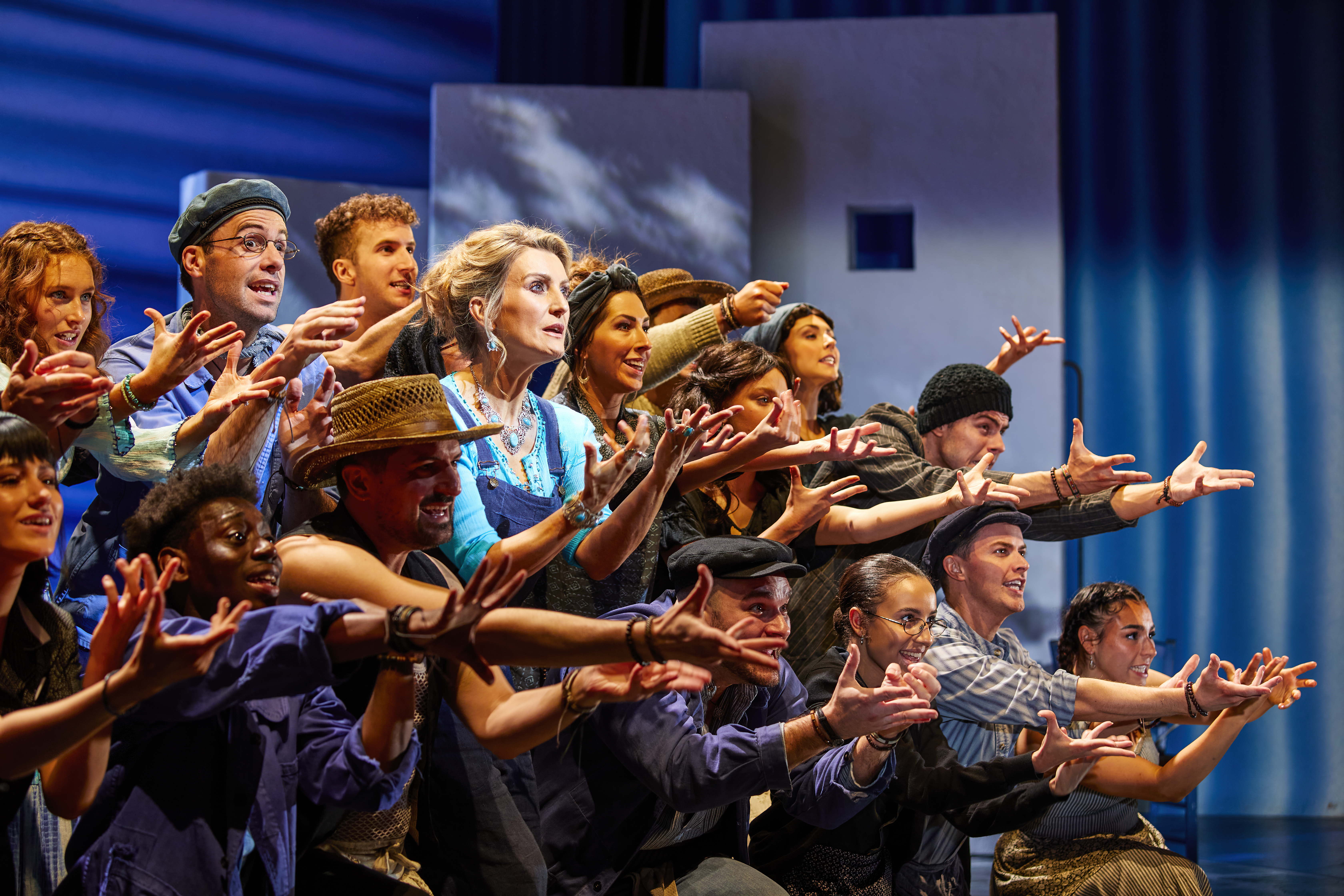 Mid shot of Sara Poyzer (centre) surrounded by the ensemble cast onstage in MAMMA MIA!