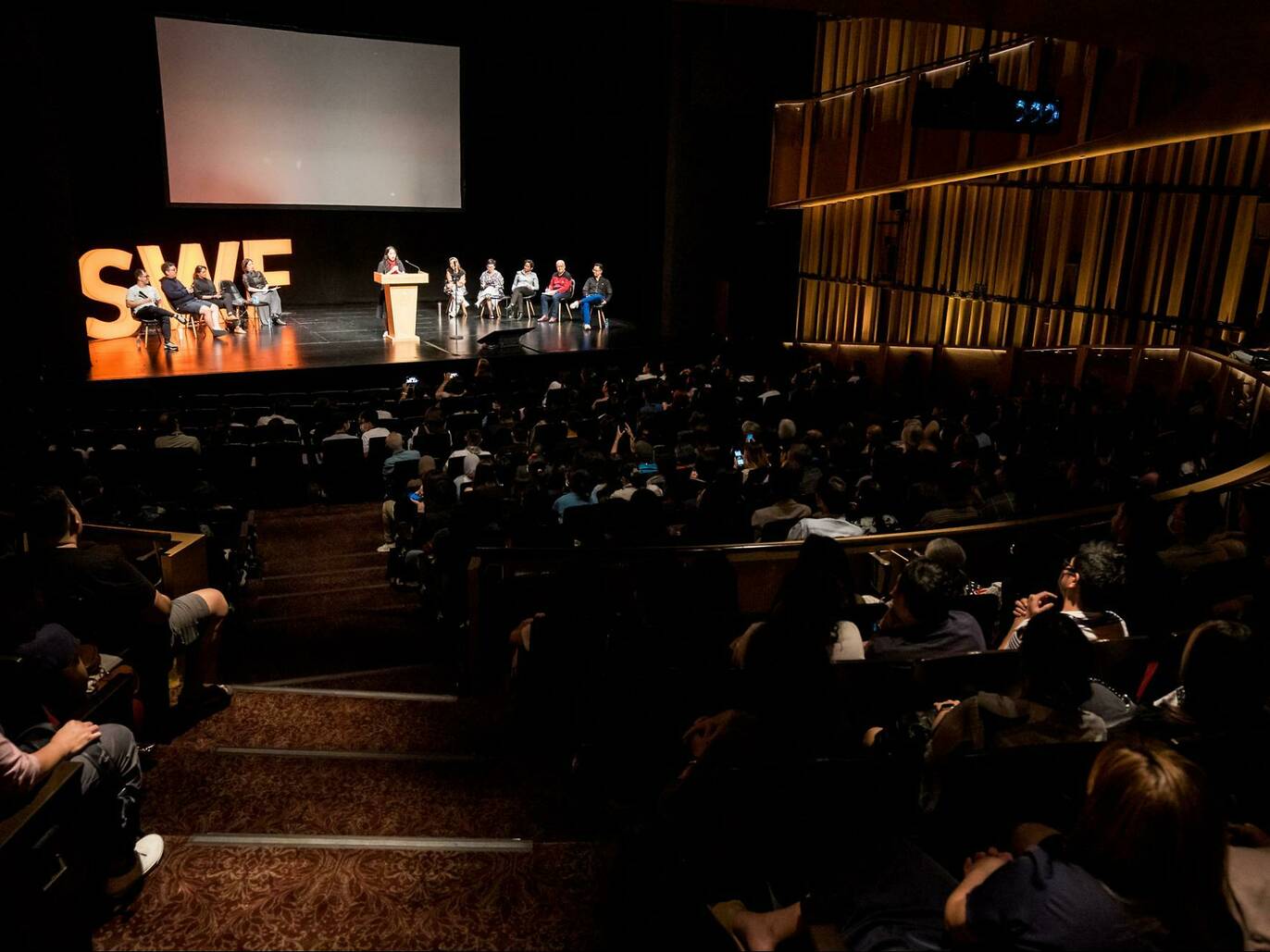 Wide shot of a crowd listening to a panel of speakers onstage 
