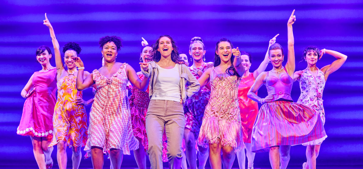 Wide shot of Tanisha Butterfield, Jess Michelmore, and Freya Humberstone (left to right) with the ensemble cast onstage in MAMMA MIA! 